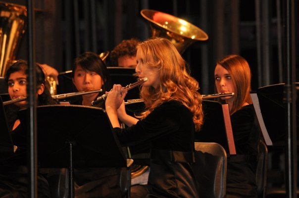 A girl, Katie, sits on a stage in a formal black dress with her blond hair styled in curls playing a flute in a band concert.