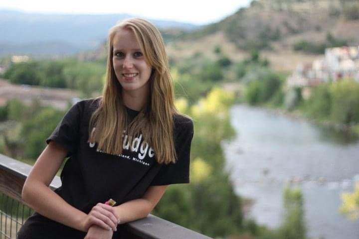 Katie stands facing the camera while smiling with a river and mountains in the background.