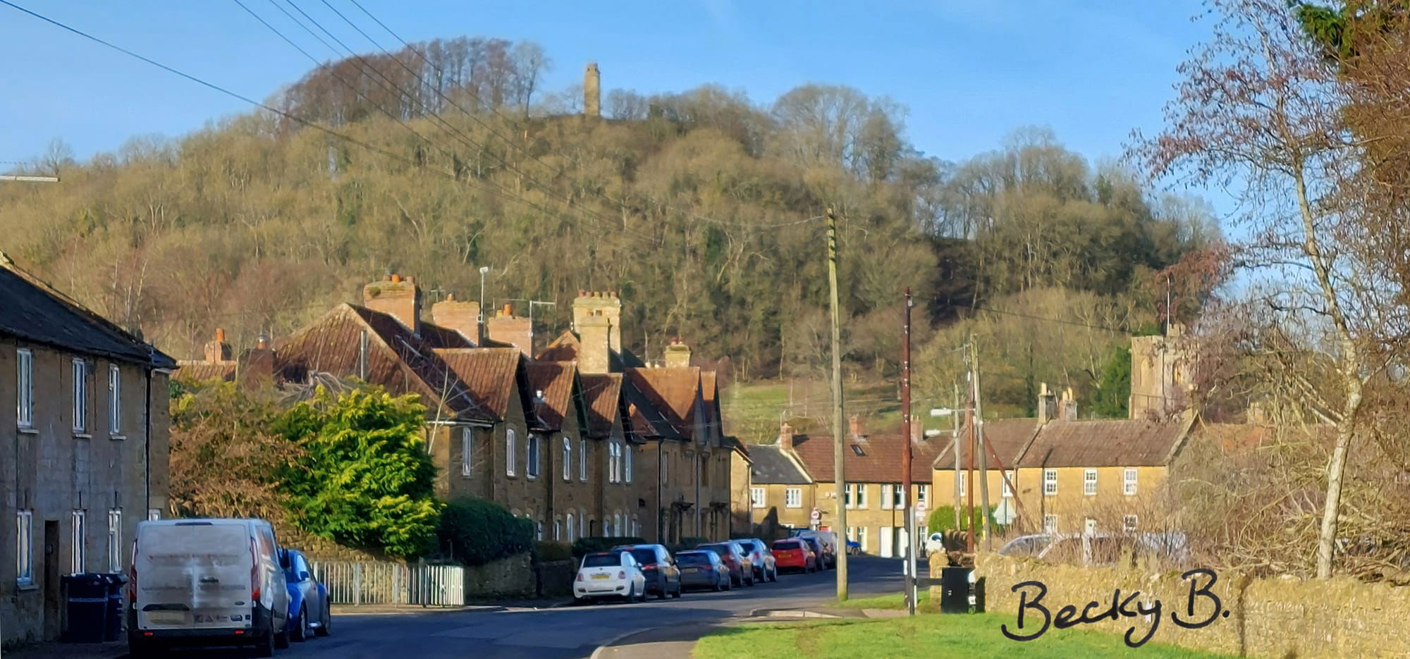 Colour photo taken looking westwards along Yeovil road from just after Montacute Garage. The left side of the road is lined with sand-coloured hamstone houses and also parked cars. Ahead (where the road bears round to the right) there are more houses and also two on the right side. Looming over the scene is St Michael’s Hill topped by the tall hamstone tower. The sky is blue and there are no leaves on the deciduous trees on the hill, giving the impression of a bright and crisp winter’s day. The photo is signed ‘Becky B’ in the bottom right corner. 