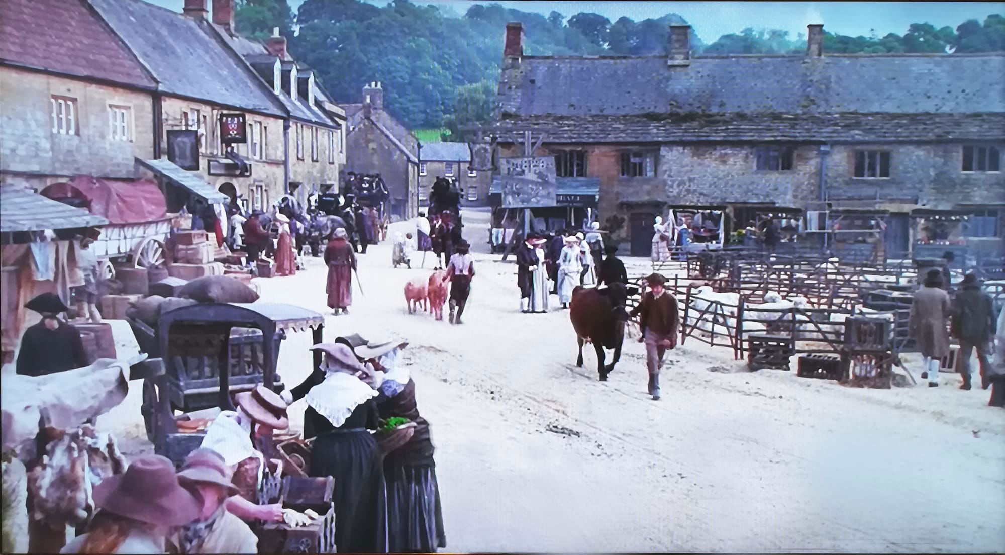 Colour photo of Montacute Borough viewed looking south with the Phelips on the left and the Milk House ahead. The picture has been screen-shotted from Ang Lee's 1995 film 'Sense and Sensibility'. There are pens containing sheep in the Borough and a man is leading a cow along the road while someone else leads 2 goats. There are stalls along the left-hand side of the road. People are browsing the stalls and standing in the Borough; dress is late 18th century. 