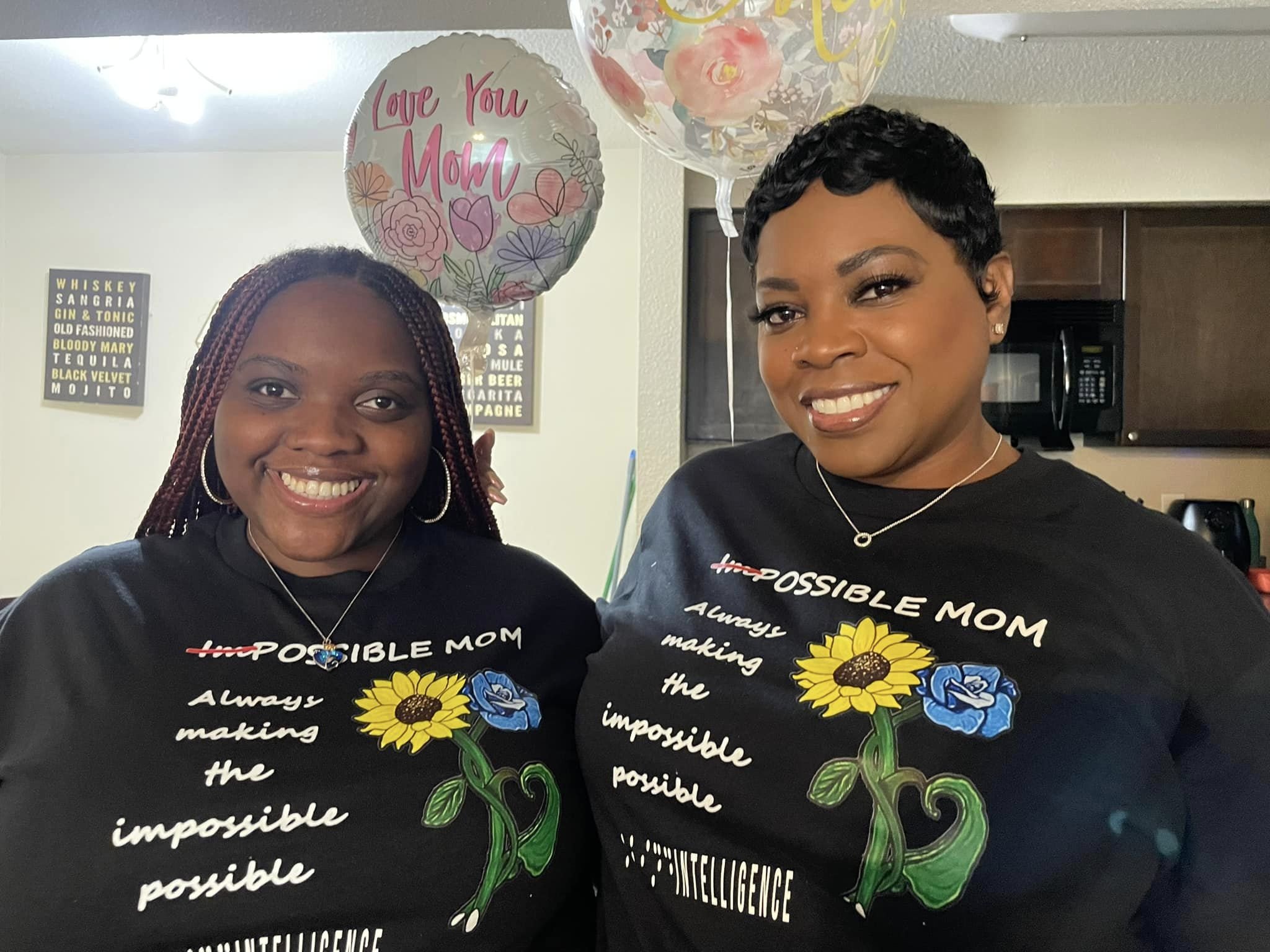 This picture is an image of Professor Blindie and and her mother standing together in a kitchen smiling, with baloons in the background the baloons say love you mom.  They are both wearing a black t-shirt with white letters that read IMPOSSOBLE MOM with the a red slash over the IM.  Always making the Impossible Possible, Blind Intelligence.  Their is Sunflower and a blue flower artwork on the side of the letters.